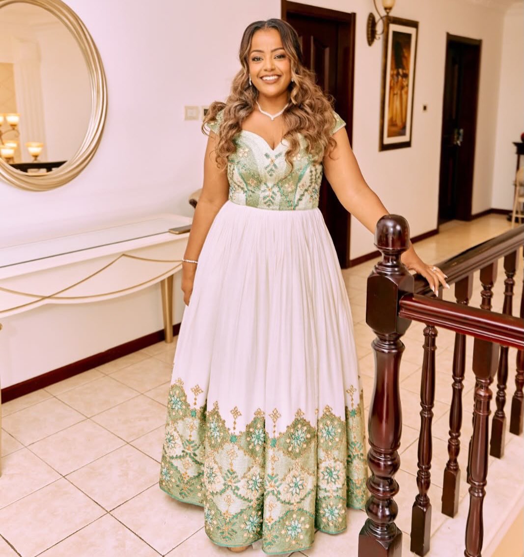 A woman wearing a formal floor-length Habesha Dress with a white skirt and green embroidered bodice and hem, standing indoors near a wooden staircase railing.
