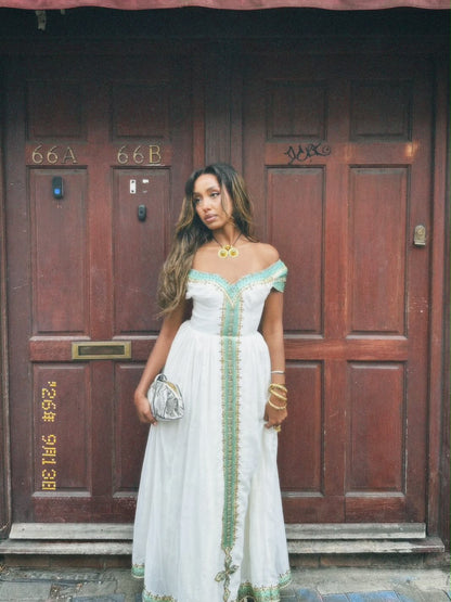 A woman stands in front of a large dark brown wooden door wearing a white off-the-shoulder Ethiopian national dresses with mint green embroidery and a central vertical embellishment. She is holding a small metallic clutch and wearing gold jewelry.