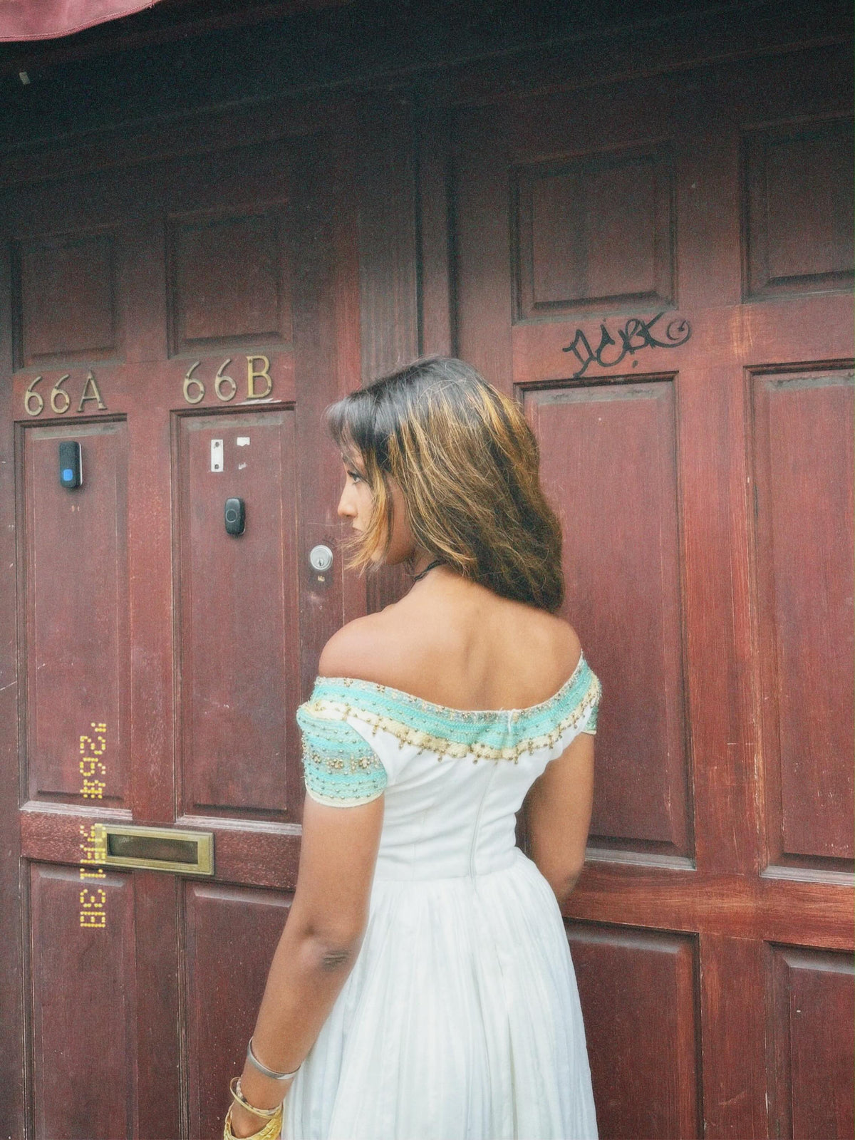 A woman wearing an off-the-shoulder white Ethiopian national dresses with a light blue and gold embroidered neckline stands in front of dark reddish-brown wooden doors.