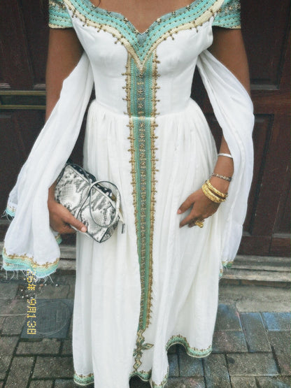A woman wearing a white maxi Ethiopian national dresses with mint green and gold embroidery, holding a snakeskin-patterned clutch bag.