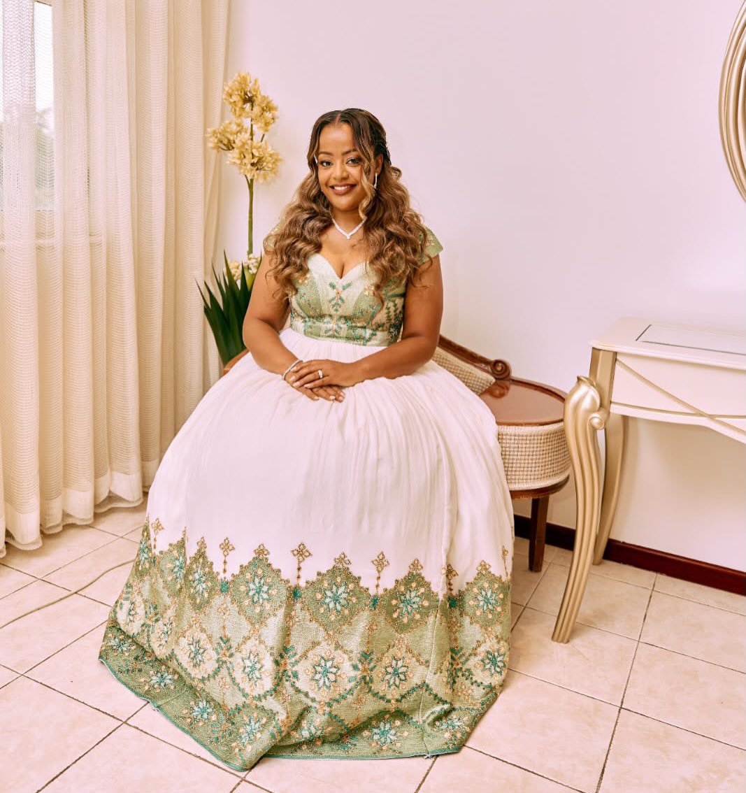 A woman wearing a formal ball Habesha Dress with a white skirt and green embroidered bodice and hem, seated in a chair in an indoor room with curtains, a console table, and a plant.