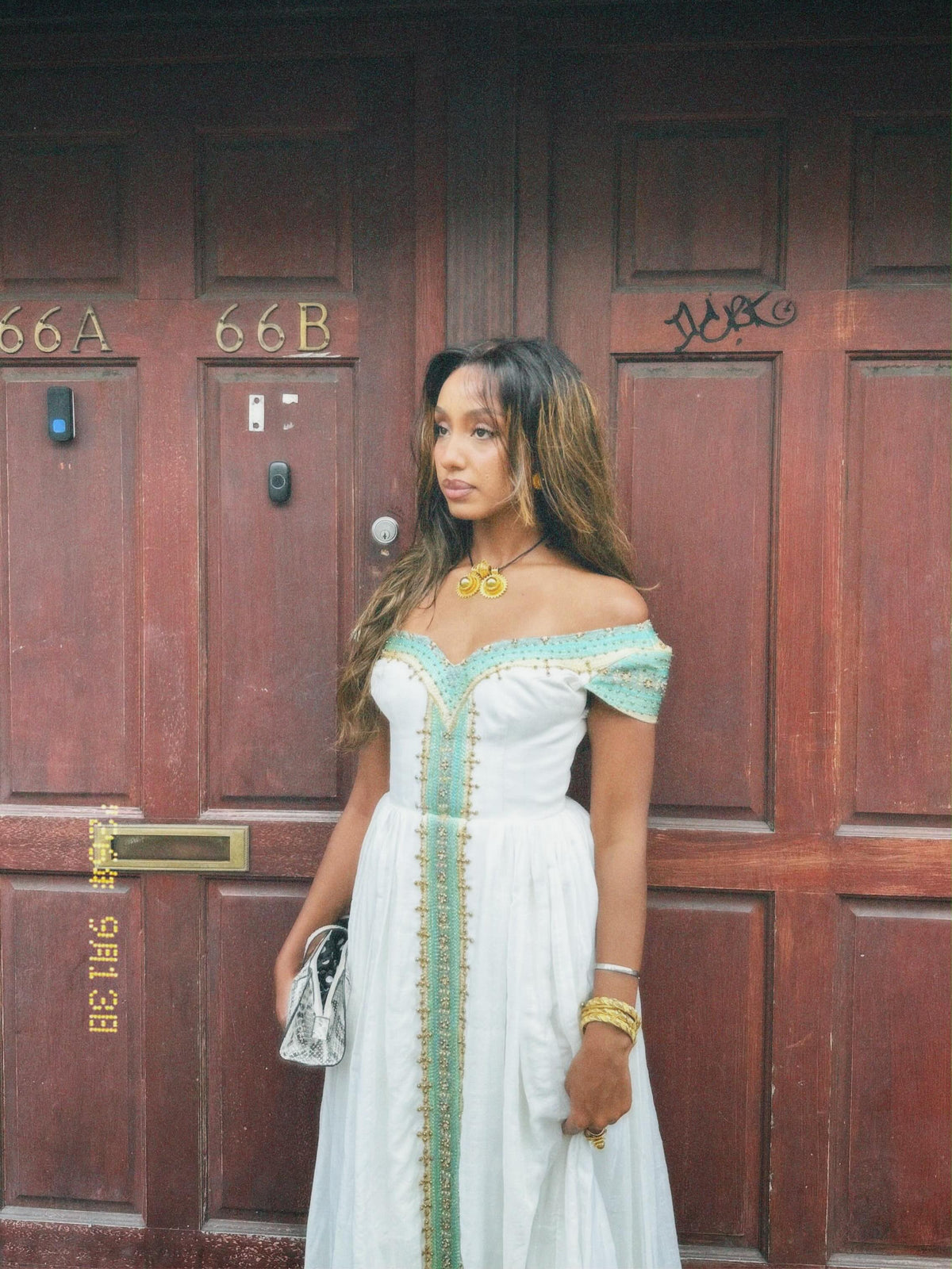 A woman wearing a white off-the-shoulder Ethiopian national dresses with mint green and gold embroidery stands in front of a dark brown wooden door, holding a small silver clutch.