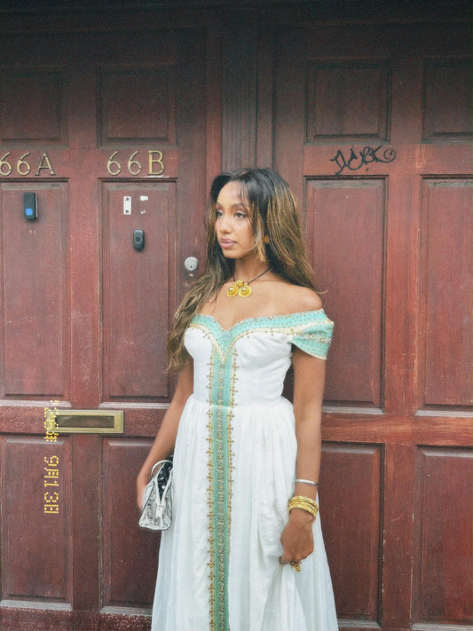 A woman wearing a white off-the-shoulder Ethiopian national dresses with mint green and gold embroidery stands in front of a dark brown wooden door, holding a small silver clutch.