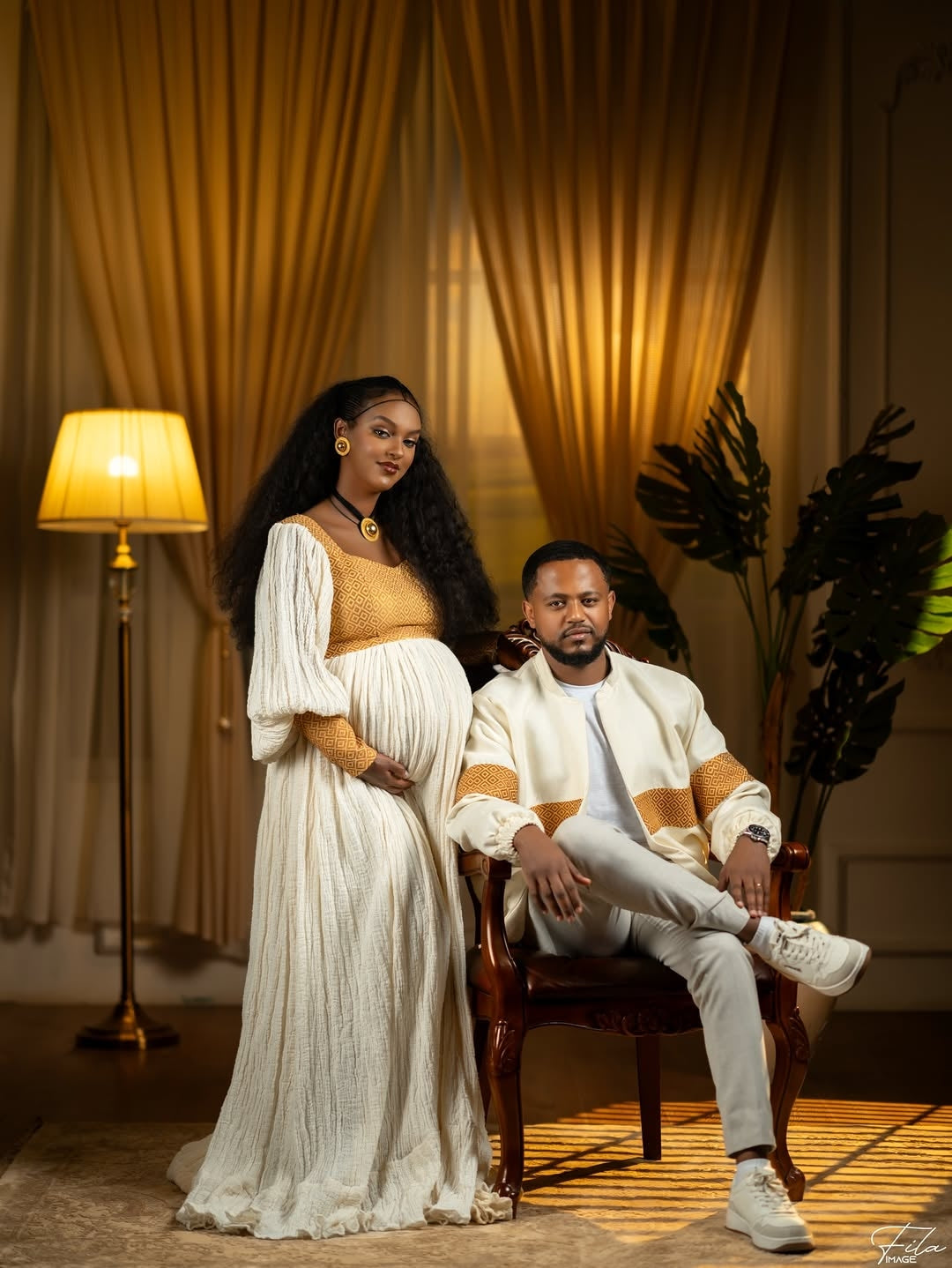 Pregnant woman in a white dress sitting next to a man in a room with warm lighting.