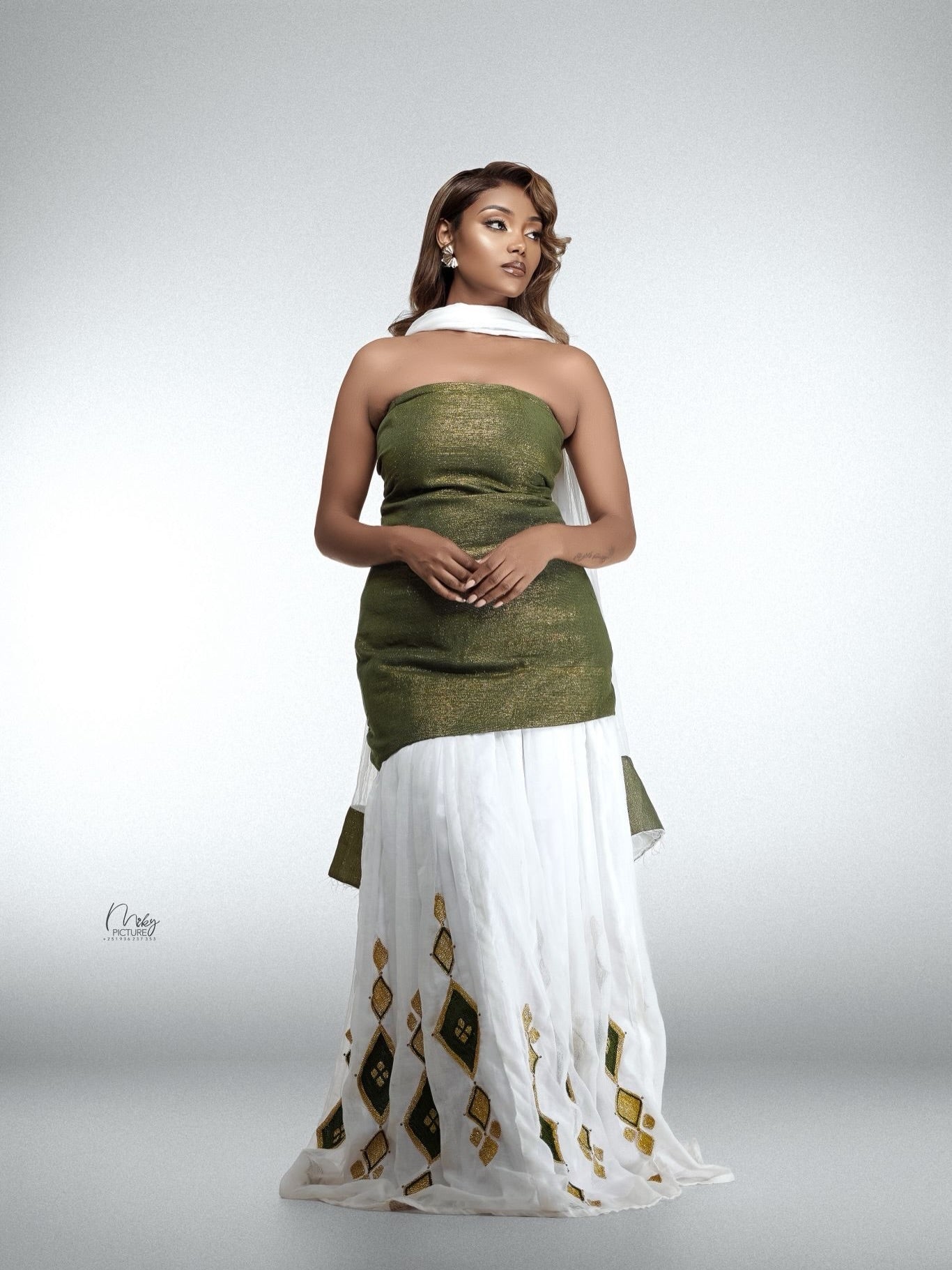 A woman modeling a traditional habesha kemis featuring an olive green strapless kurta paired with a white skirt decorated with gold geometric patterns and a white dupatta, set against a plain light gray studio background.