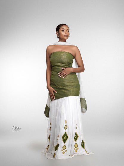 A model wearing a traditional habesha kemisconsisting of an olive green strapless kurta, a white skirt with green and gold diamond patterns, and a white dupatta, set against a light gray studio background.