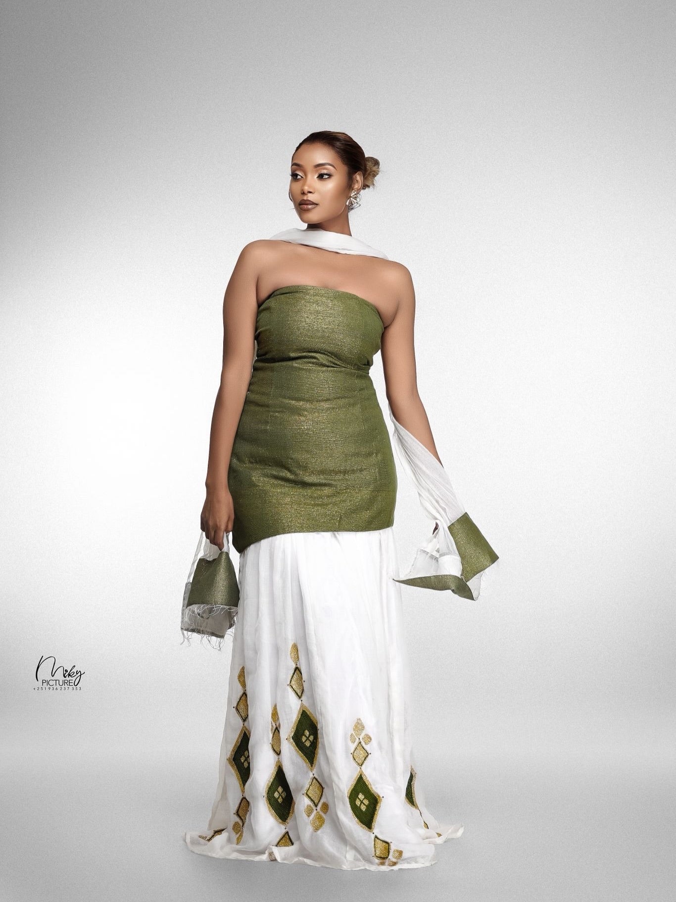 A woman models a strapless two-piece dress with a textured olive green top and a flowing white habesha kemis featuring green and gold diamond patterns. She is posed against a neutral gray studio background, holding a matching green clutch.