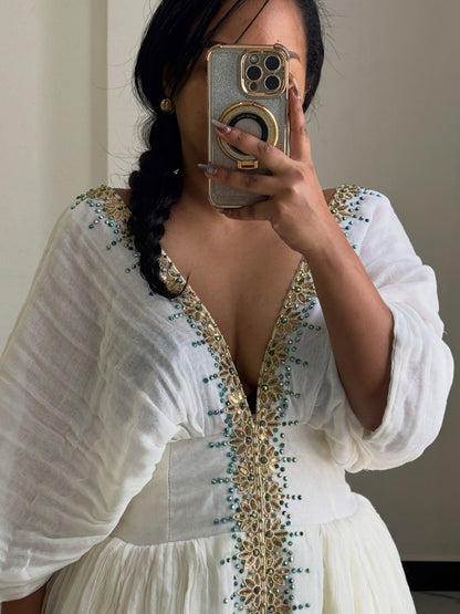 A woman takes a mirror selfie wearing a white long-sleeve Habesha Dress with gold and teal beaded embroidery along the deep V-neckline and waistband.