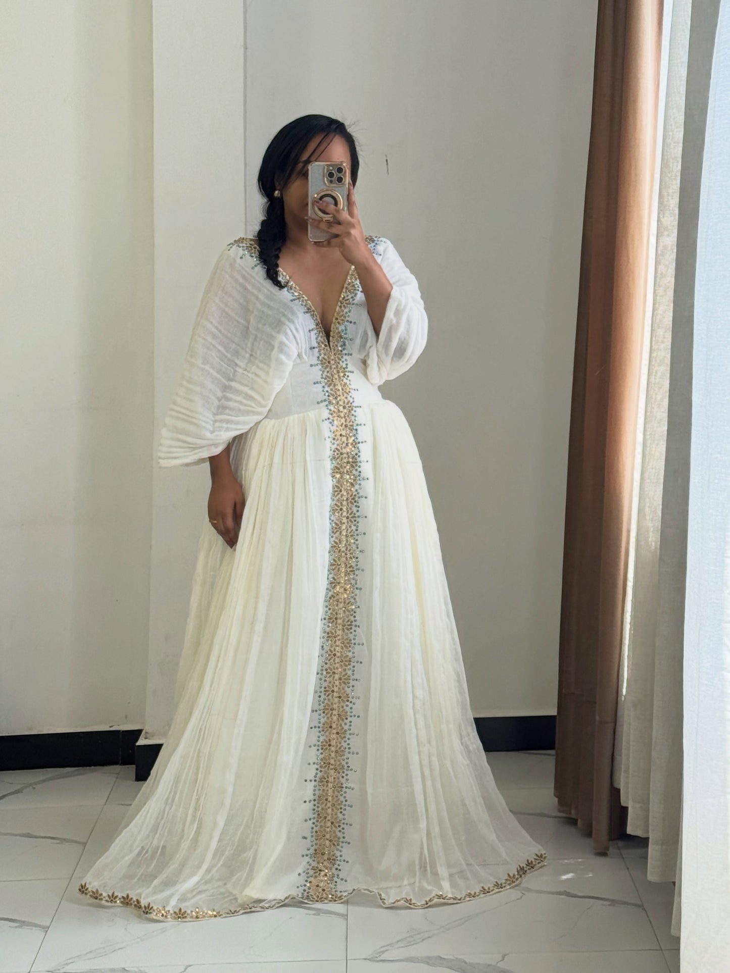 A woman takes a mirror selfie wearing a long white Habesha Dress with gold embroidery down the front and on the hem, set against an indoor background with curtains.
