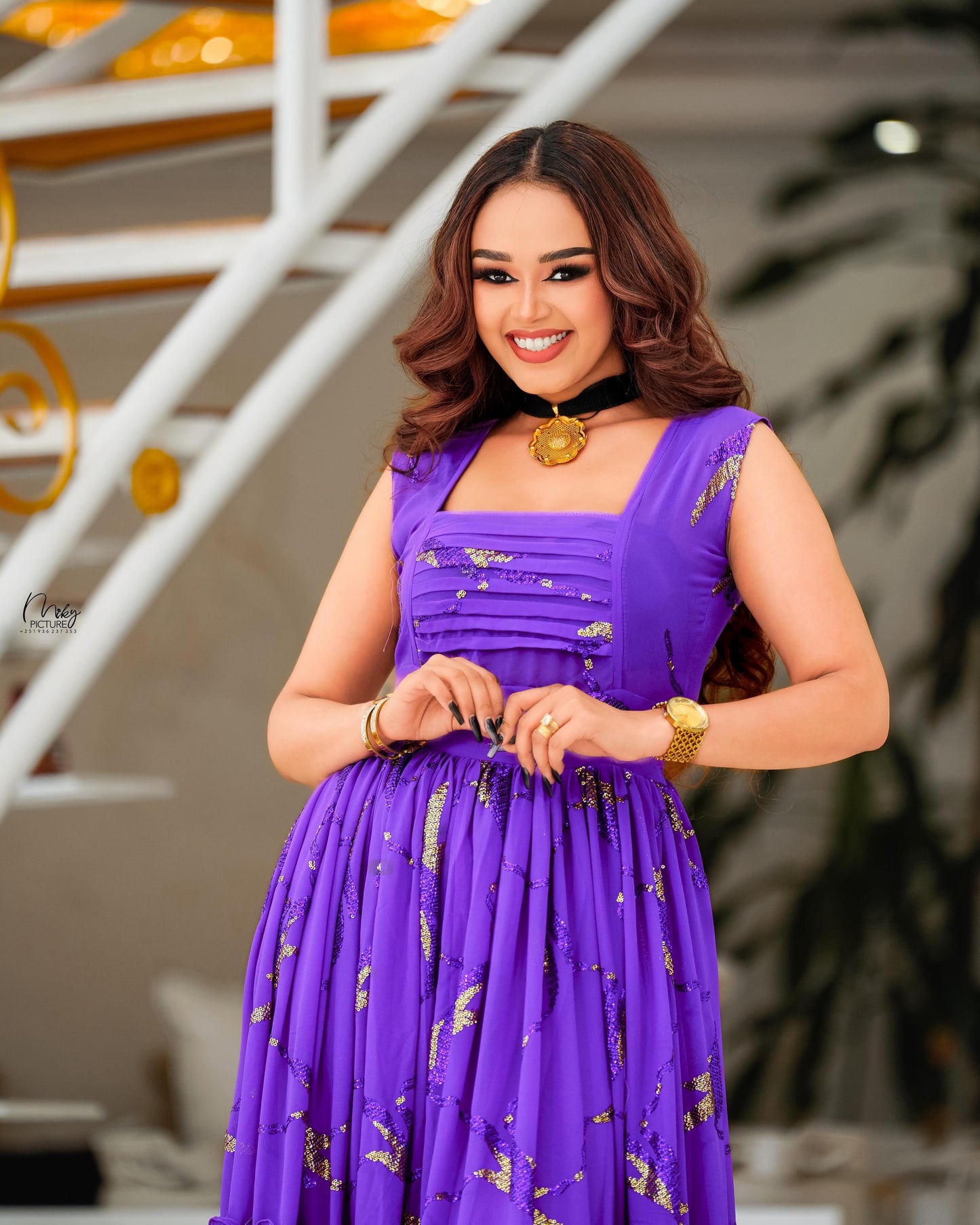 Woman in a purple habesha dress with gold accents standing indoors.