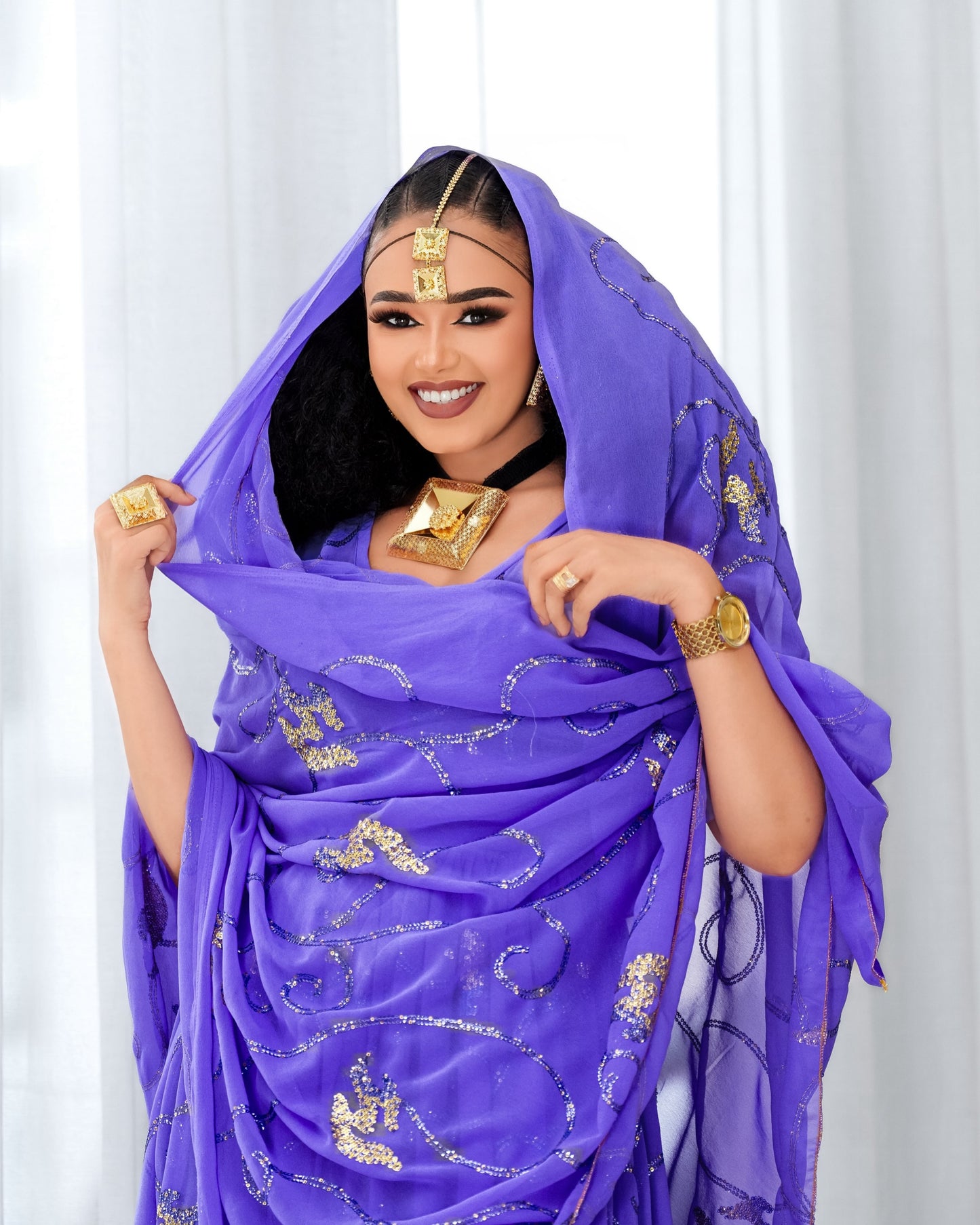 A woman wearing a vibrant purple traditional Habesha Dress with gold patterns, holding the fabric outwards to display its drape and design. She is accessorized with gold jewelry, including a headpiece, necklace, rings, and a watch, set against a soft white curtain background.