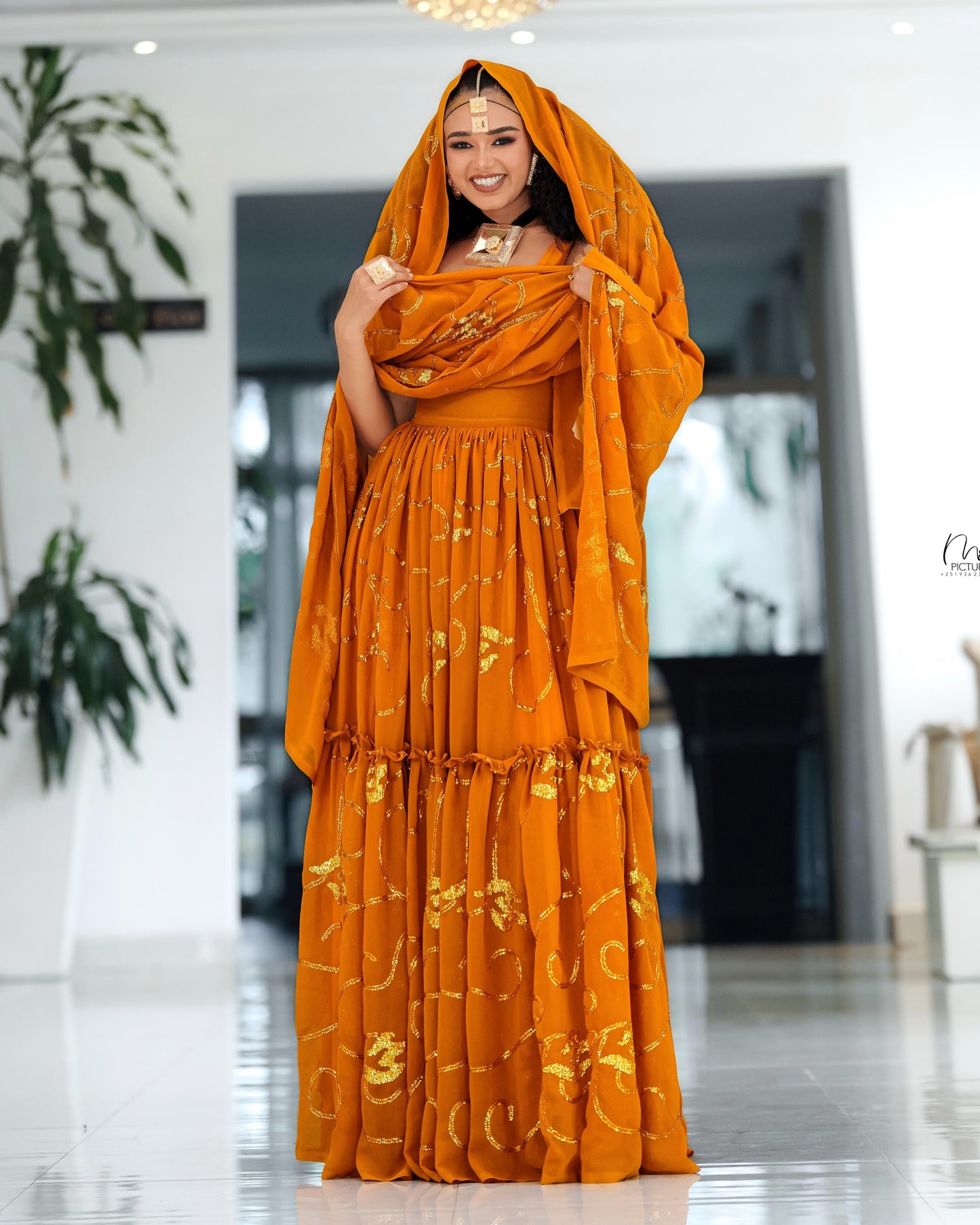 A woman wearing a bright orange traditional Habesha Chiffon  with gold embroidery and a matching draped headscarf, standing in a bright indoor showroom.