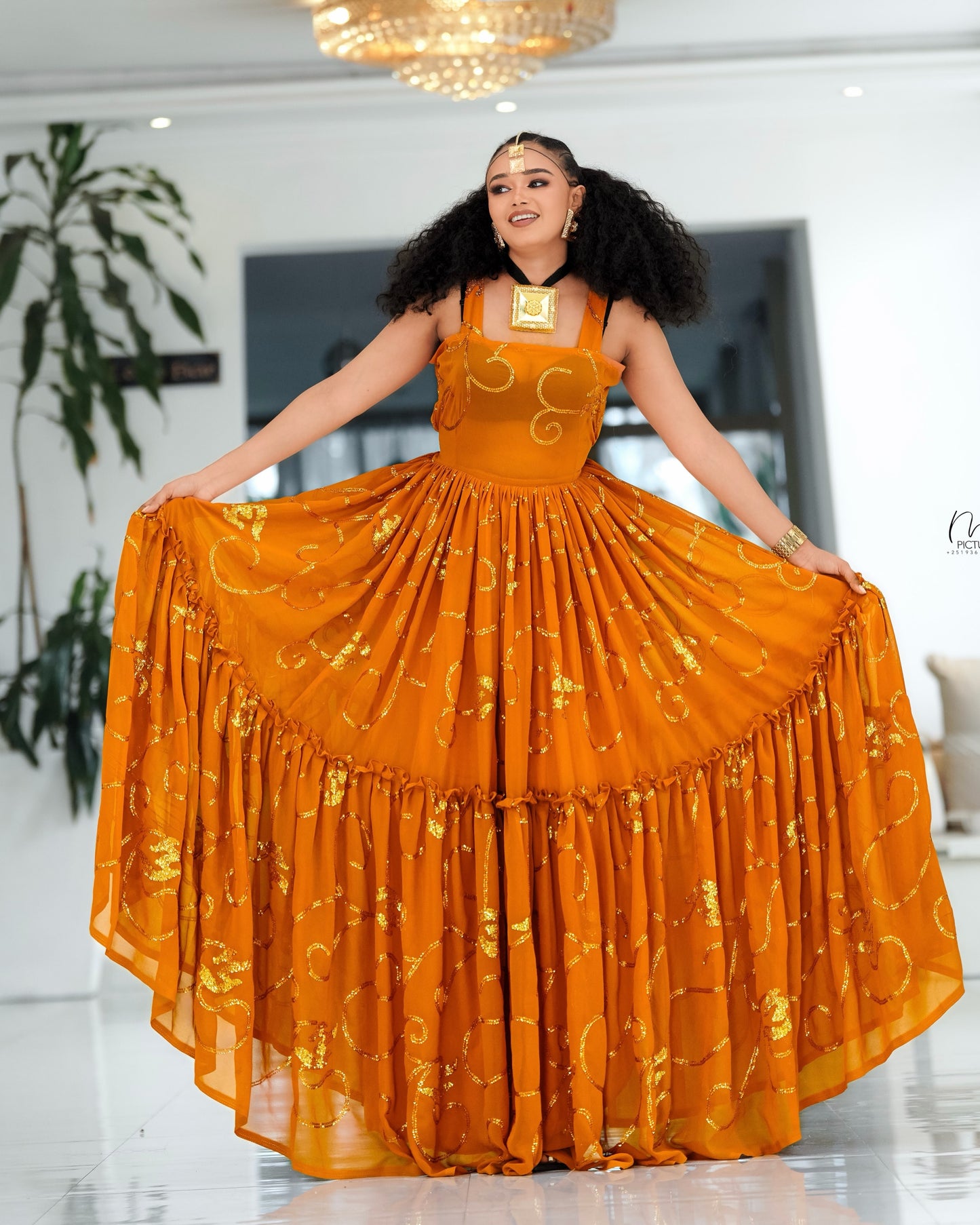 A woman modeling a voluminous orange traditional Habesha Chiffon  with gold embroidery, holding out the wide skirt in a bright indoor setting.
