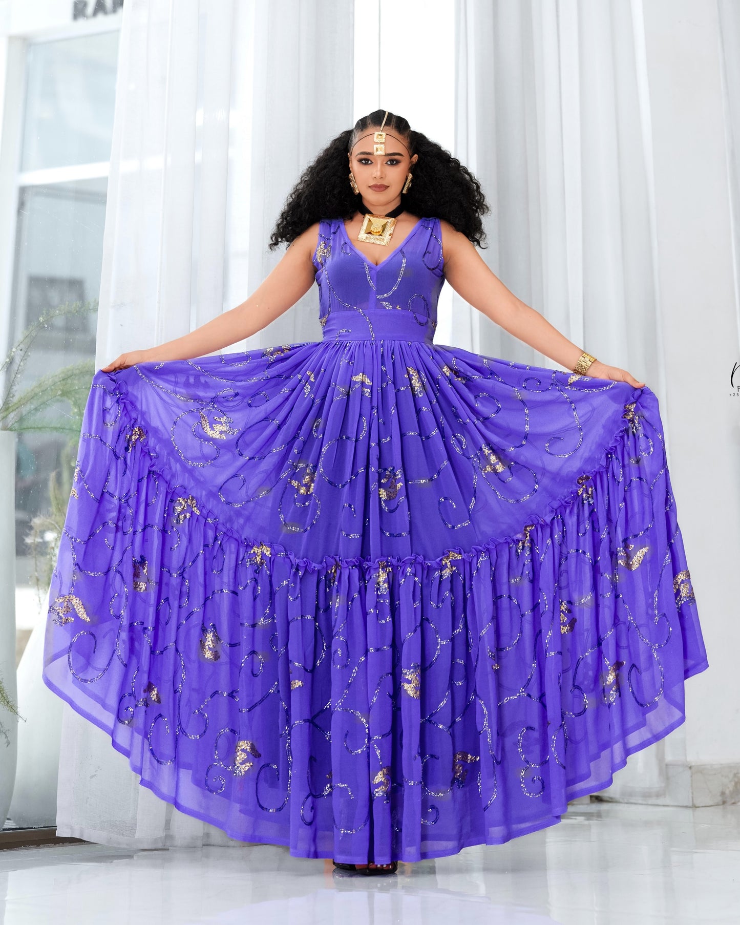 A woman modeling a voluminous purple sleeveless Habesha Dress with gold floral embroidery, holding out the wide skirt against a bright white curtain background.