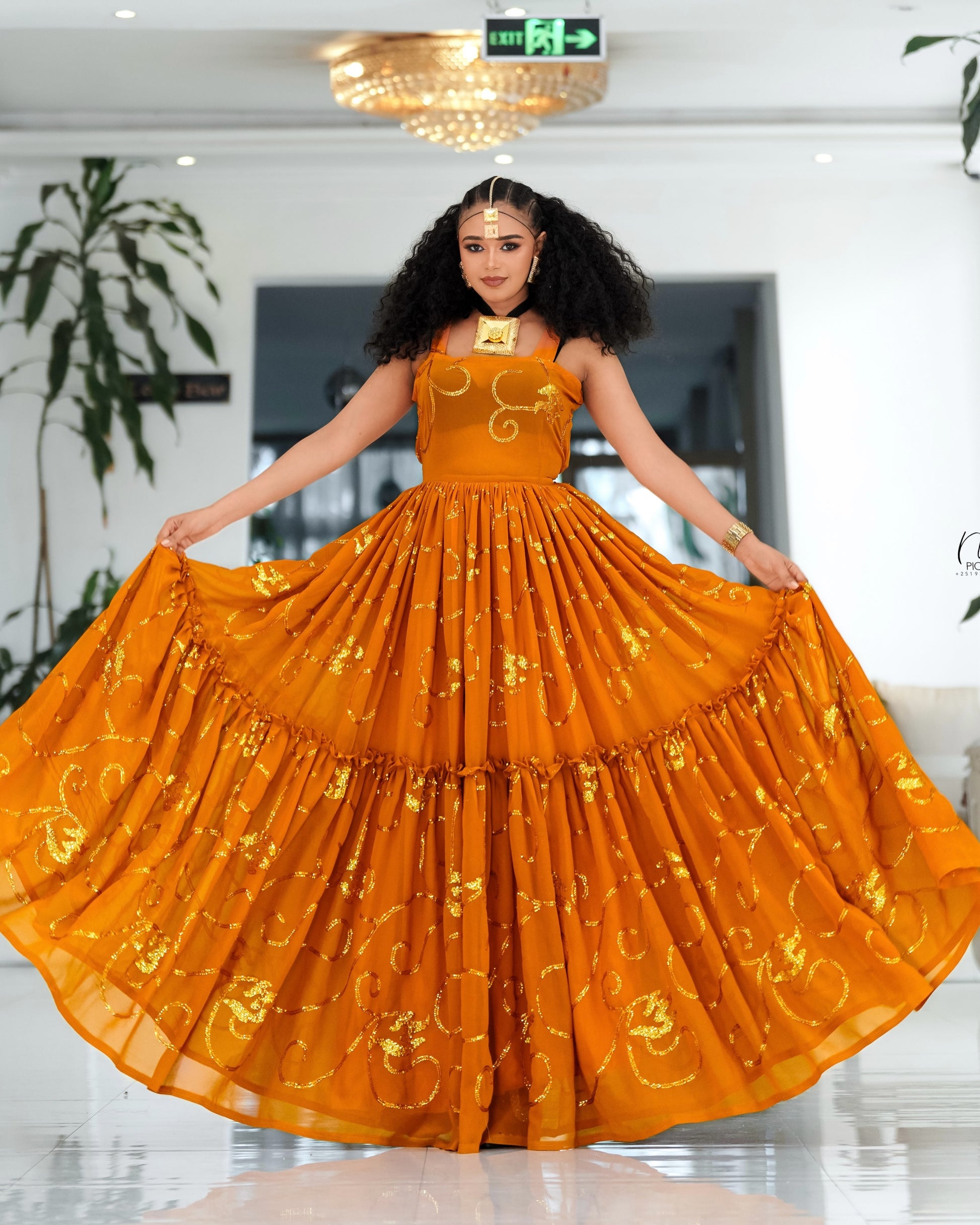 A woman modeling a sleeveless, tiered orange-gold Habesha Chiffon  with gold floral embroidery, holding out the wide skirt in an indoor setting.