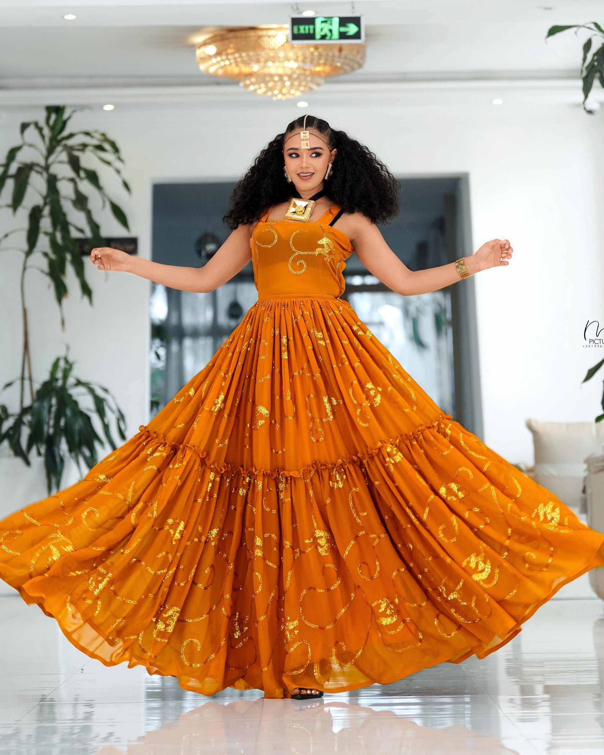 A woman wearing a vibrant orange sleeveless maxi Habesha Chiffon  with gold floral patterns, twirling in a spacious, bright white indoor space with plants and a chandelier.