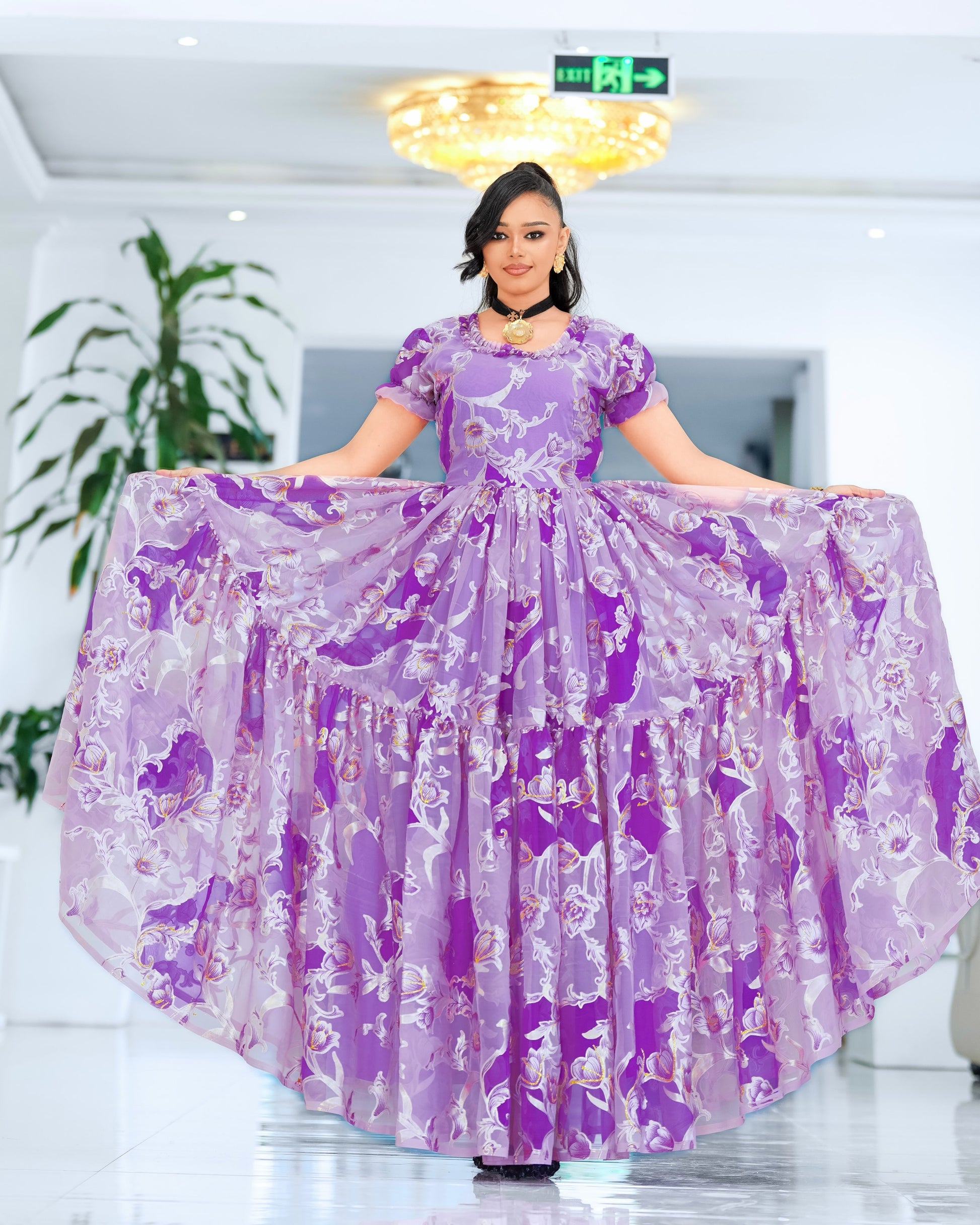 A woman stands indoors holding out a voluminous lavender and purple  Ethiopian chiffon with short puff sleeves and a tiered skirt. The background shows a bright white hallway with a chandelier, plant, and exit sign.