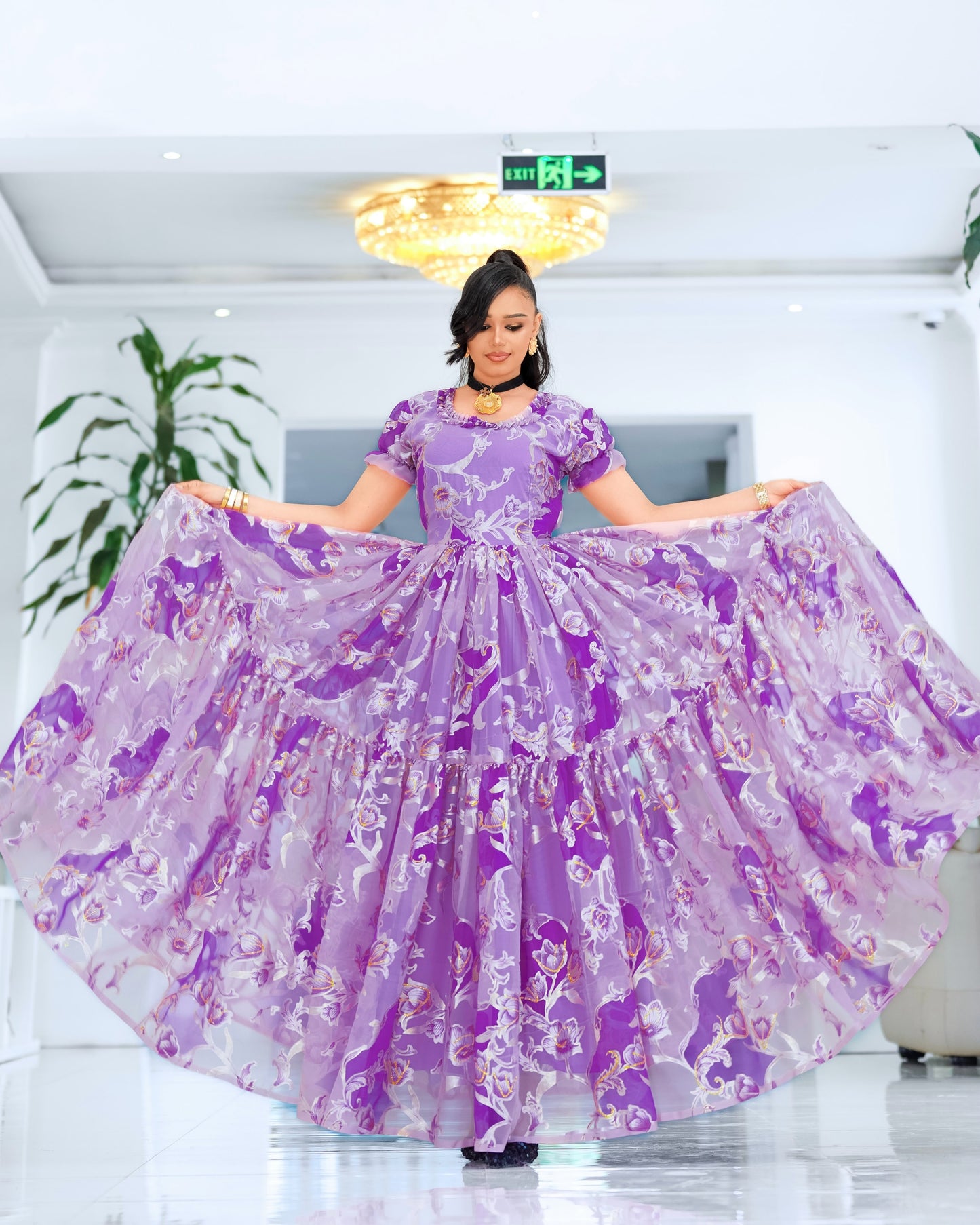 A woman stands indoors holding out a voluminous lavender and purple patterned  Ethiopian chiffon with short sleeves and a tiered skirt. The dress has a floral or abstract print in shades of purple, pink, and white.