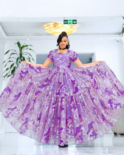 A woman stands indoors holding out a voluminous lavender and purple patterned  Ethiopian chiffon with short sleeves and a tiered skirt. The dress has a floral or abstract print in shades of purple, pink, and white.