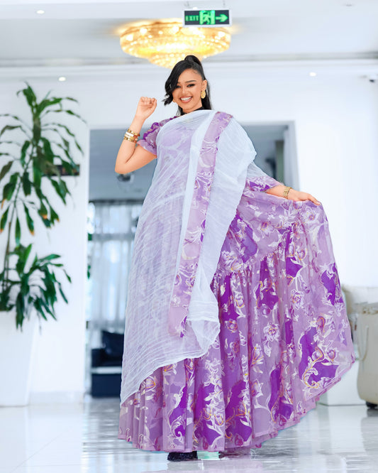 A woman modeling a traditional Ethiopian chiffon  featuring a purple and lavender floral printed skirt with a white dupatta draped over her shoulders, set in a bright, modern indoor hallway.