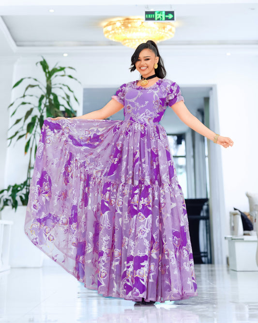 A woman wearing a purple and lavender floral print maxi Ethiopian chiffon  with short puff sleeves, twilly waist, and a full tiered skirt, posing in a bright indoor showroom.