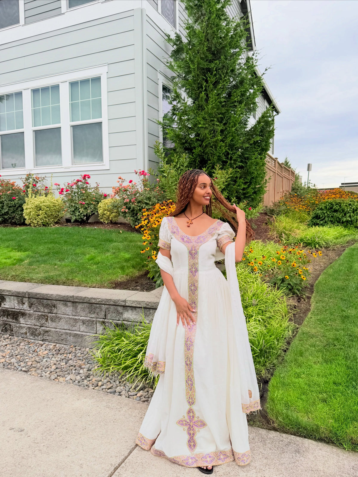 A woman wearing a white traditional Ethiopian traditional clothing with pink and gold embroidery stands on a concrete path in a landscaped garden in front of a house.