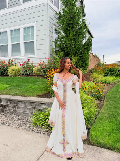 A woman wearing a white traditional Ethiopian traditional clothing with pink and gold embroidery stands on a concrete path in a landscaped garden in front of a house.