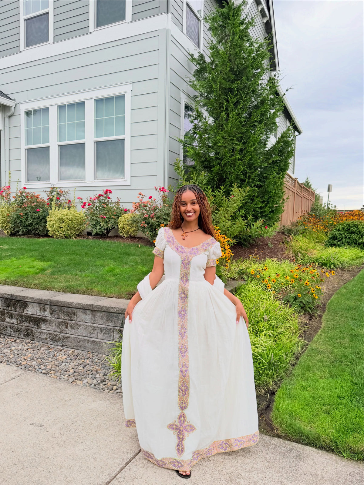 A woman wearing a long white Ethiopian traditional clothing with pink and gold embroidered details stands in a front yard in front of a light gray house.