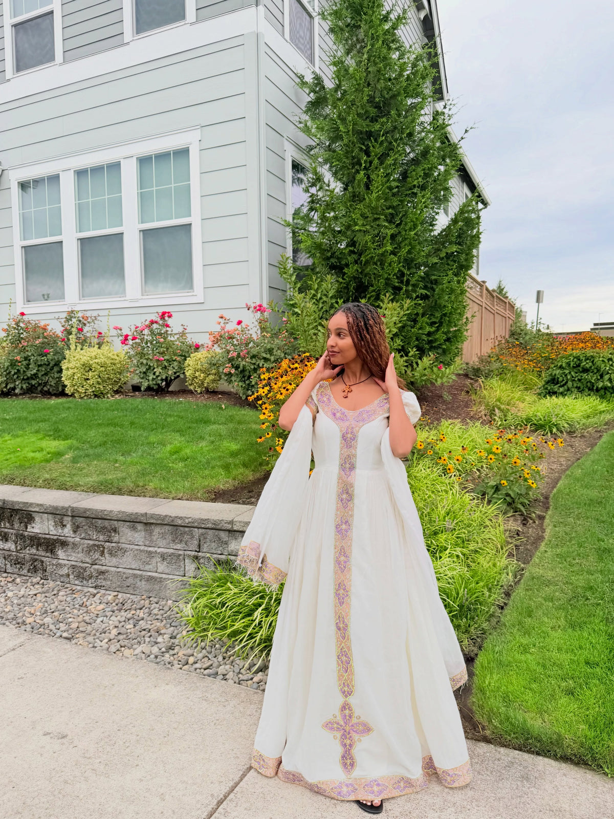 A woman wearing a long white Ethiopian traditional clothing with pink and gold embroidery, standing in a landscaped garden in front of a house.