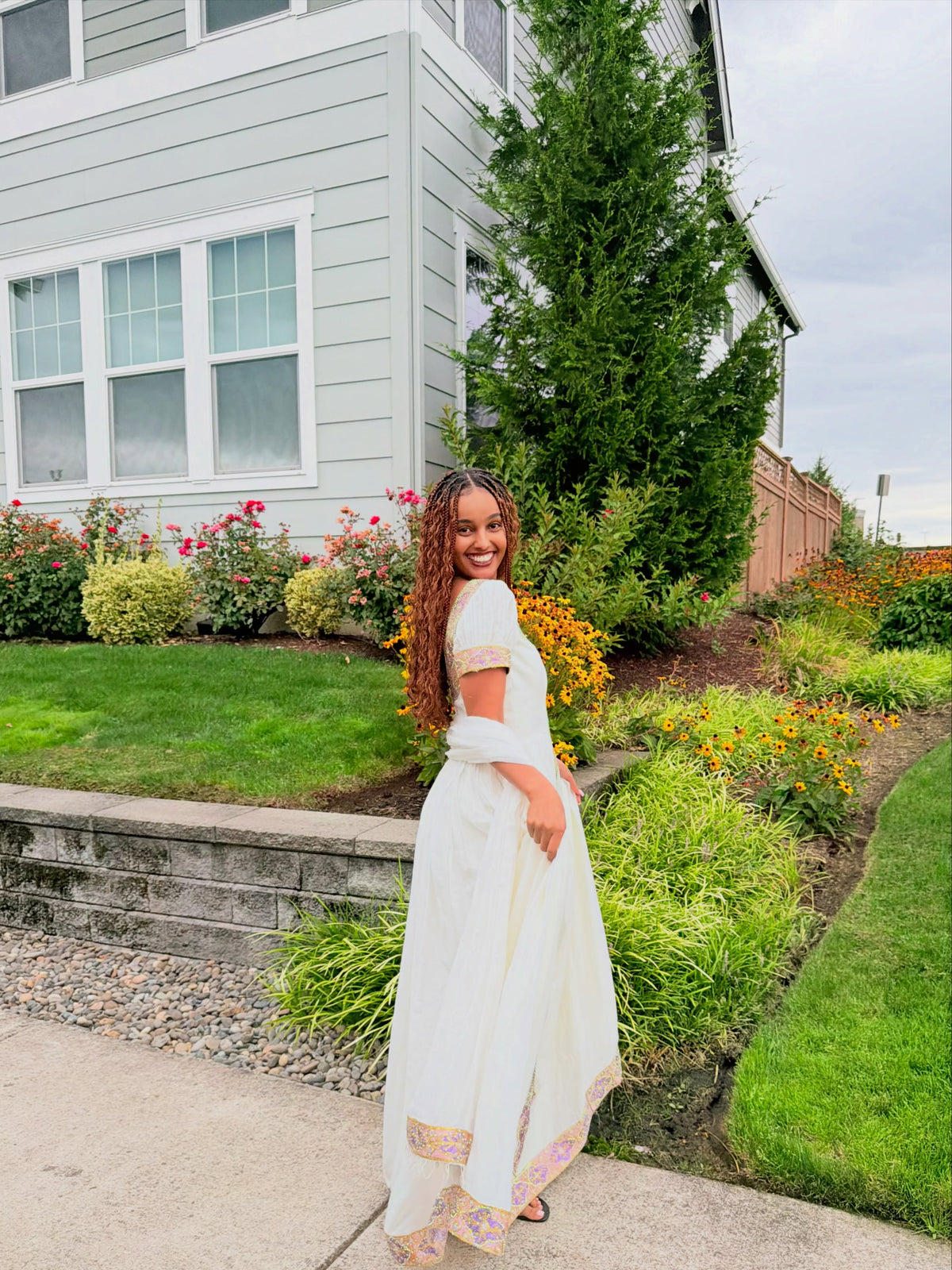 A woman wearing a white maxi Ethiopian traditional clothing with colorful embroidered borders stands in a backyard garden next to a light gray house.