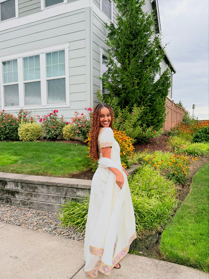 A woman wearing a white maxi Ethiopian traditional clothing with colorful embroidered borders stands in a backyard garden next to a light gray house.
