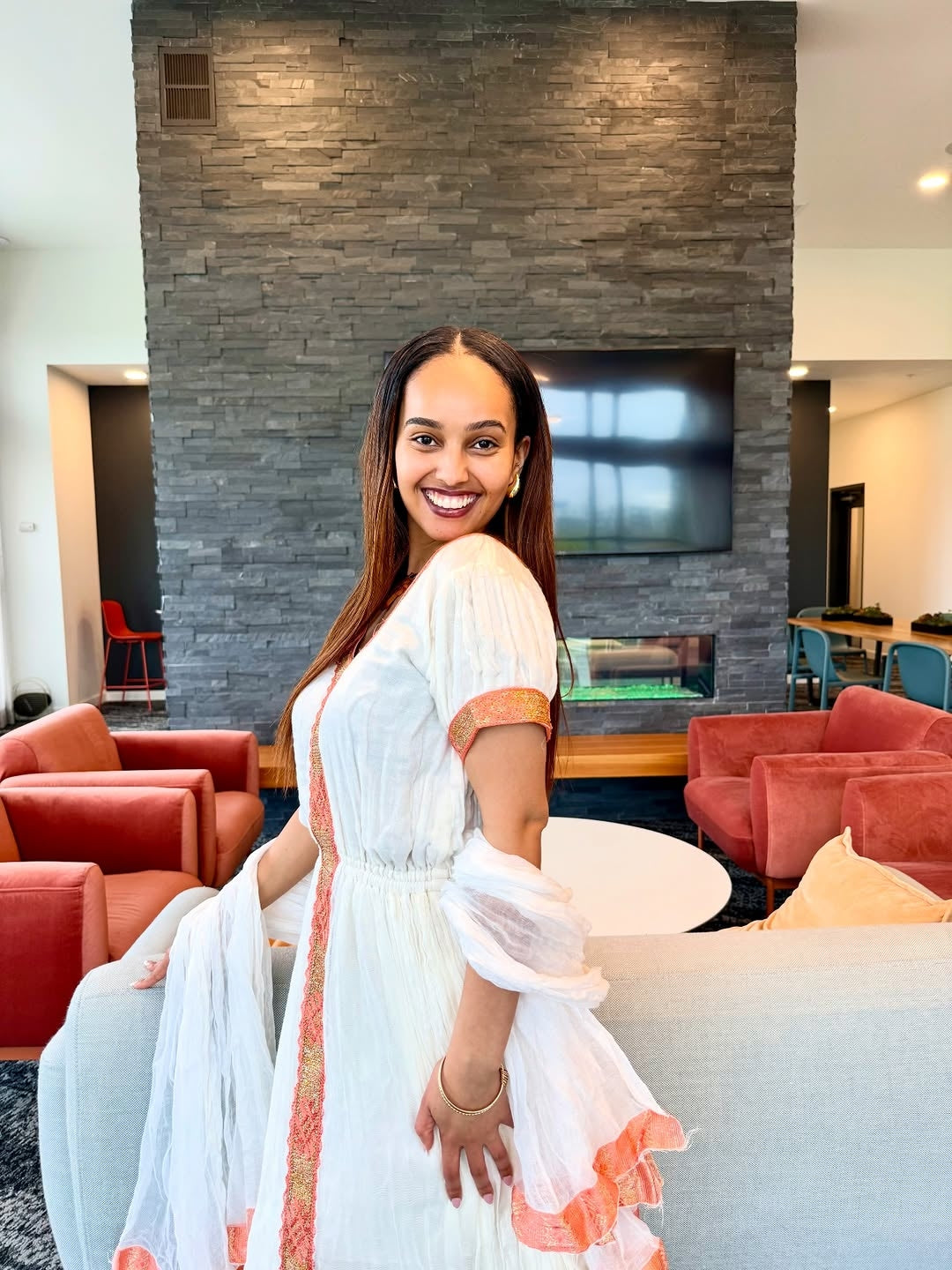 Woman in a white habesha dress  with orange accents sitting on a couch in a modern living room.