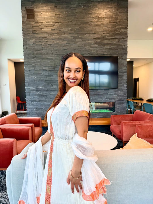 Woman in a white habesha dress  with orange accents sitting on a couch in a modern living room.