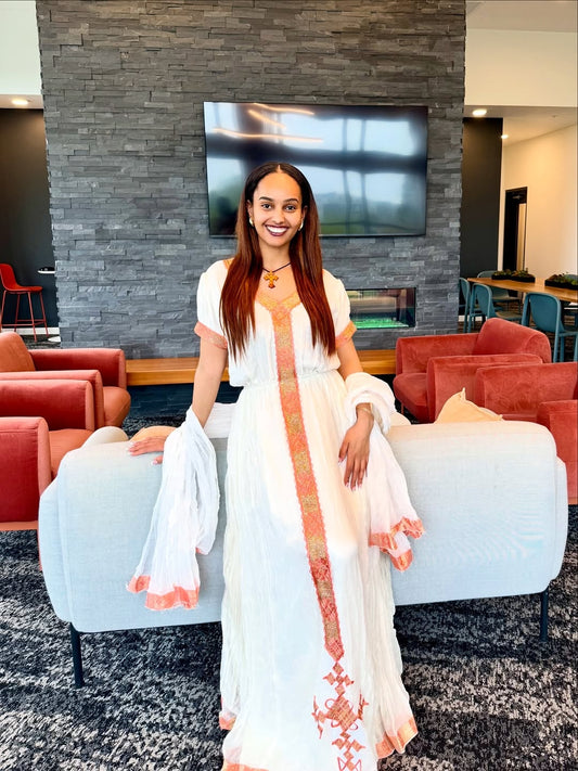 Woman in a white habesha dress  with orange accents sitting on a white chair in a modern interior setting.