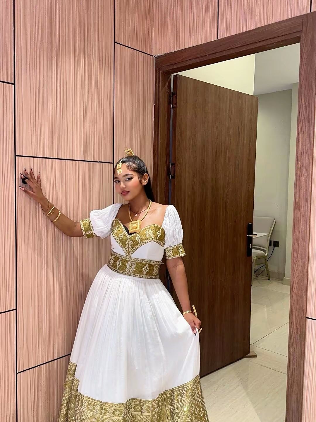 A woman wearing a traditional white and gold ethiopian dress with ornate gold embroidery, standing in an indoor setting with wood-paneled walls and a wooden door.