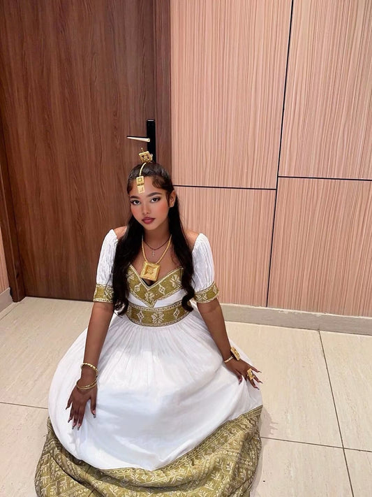 A woman wearing a traditional white and gold ethiopian dress with gold jewelry sits on a tiled floor in front of a wooden paneled wall.