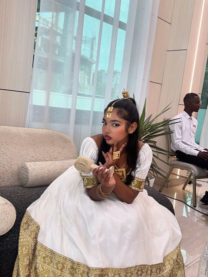 A woman wearing a traditional white ethiopian dress with gold patterns and gold jewelry sits on a sofa in a modern indoor setting, holding a small handheld fan. A man in a white shirt is seated in the background near a window.