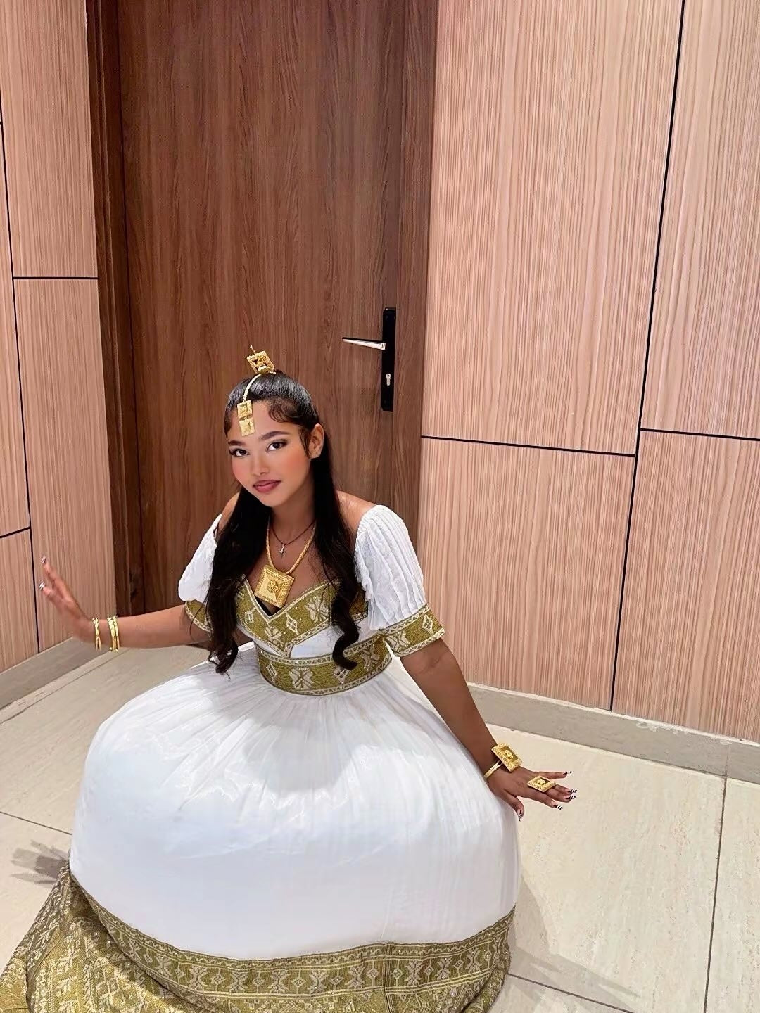 A woman wearing a traditional royal-style ethiopian dress with ornate gold embroidery, gold jewelry, and a gold headpiece, posing indoors in front of a wooden door and paneled wall.