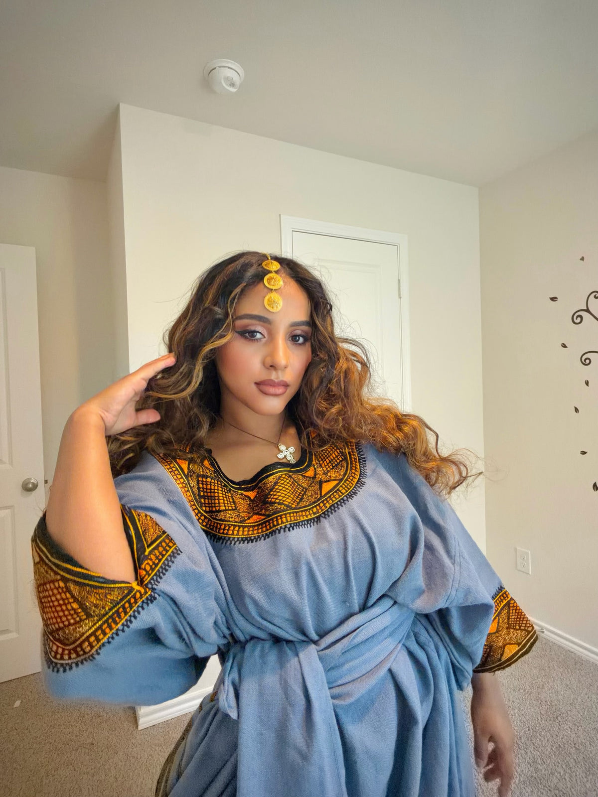 A woman wearing a light blue Ethiopian store with yellow and black patterned collar and sleeve cuffs, posing in a home interior.