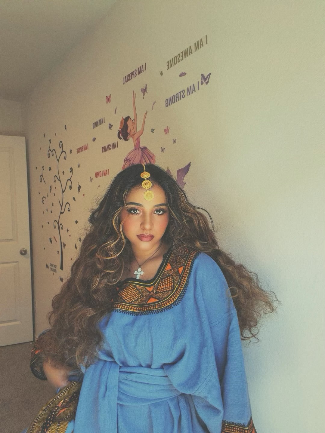 A woman with long, wavy brown hair wearing a blue Ethiopian store with an ornate gold and brown embroidered collar, standing in a room with a beige wall decorated with whimsical fairy decals.