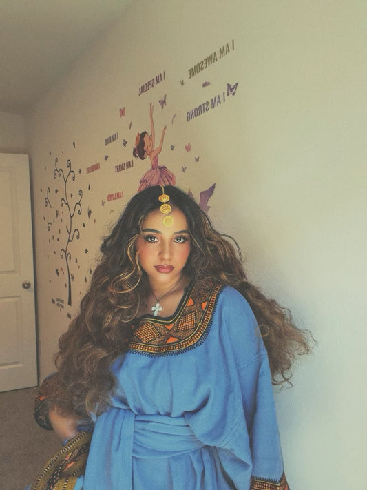 A woman with long, wavy brown hair wearing a blue Ethiopian store with an ornate gold and brown embroidered collar, standing in a room with a beige wall decorated with whimsical fairy decals.