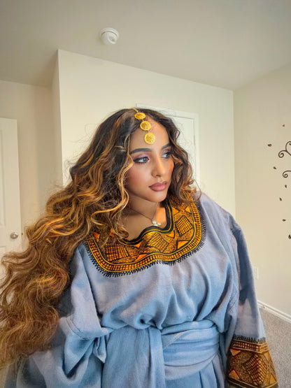 A woman wearing a light blue traditional Ethiopian store with a wide embroidered collar in orange, black, and green, styled with a gold headpiece, standing in a home interior.
