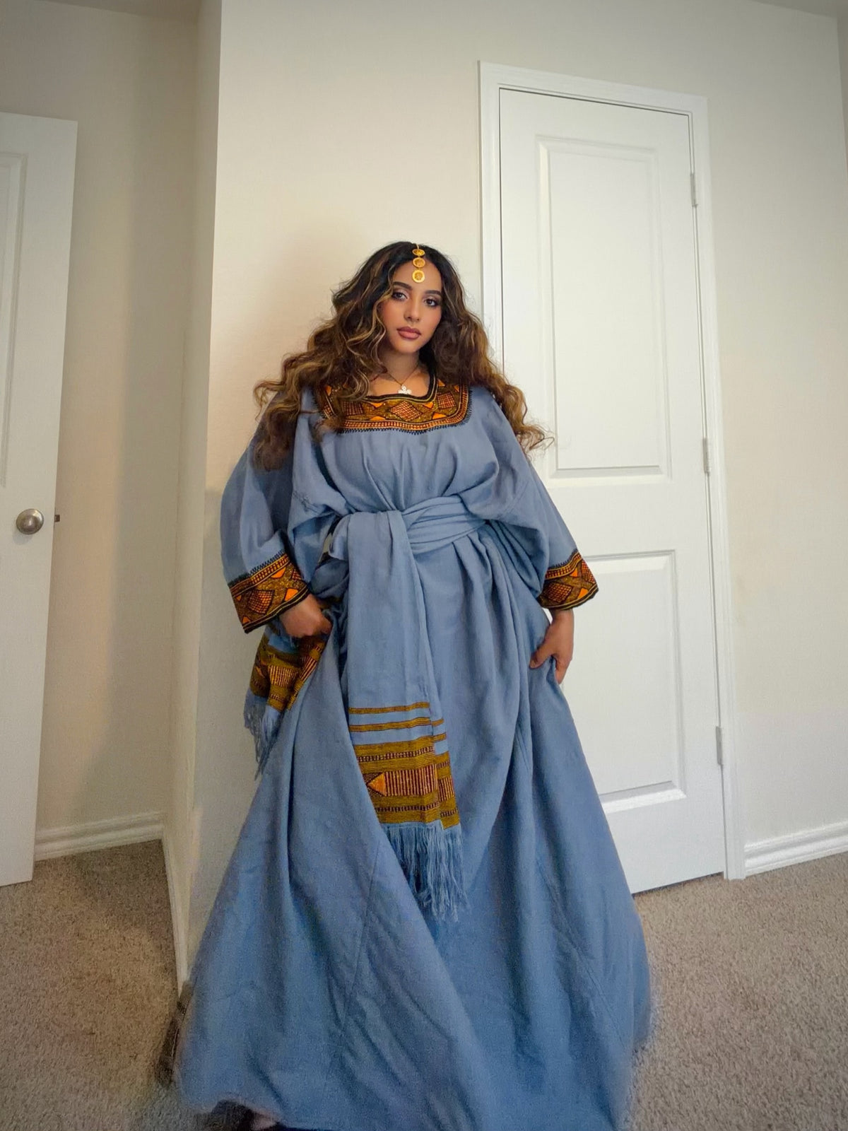 A woman wearing a light blue ancient-style costume Ethiopian store with gold and brown embroidered details, standing in a home hallway with white doors and beige carpet.
