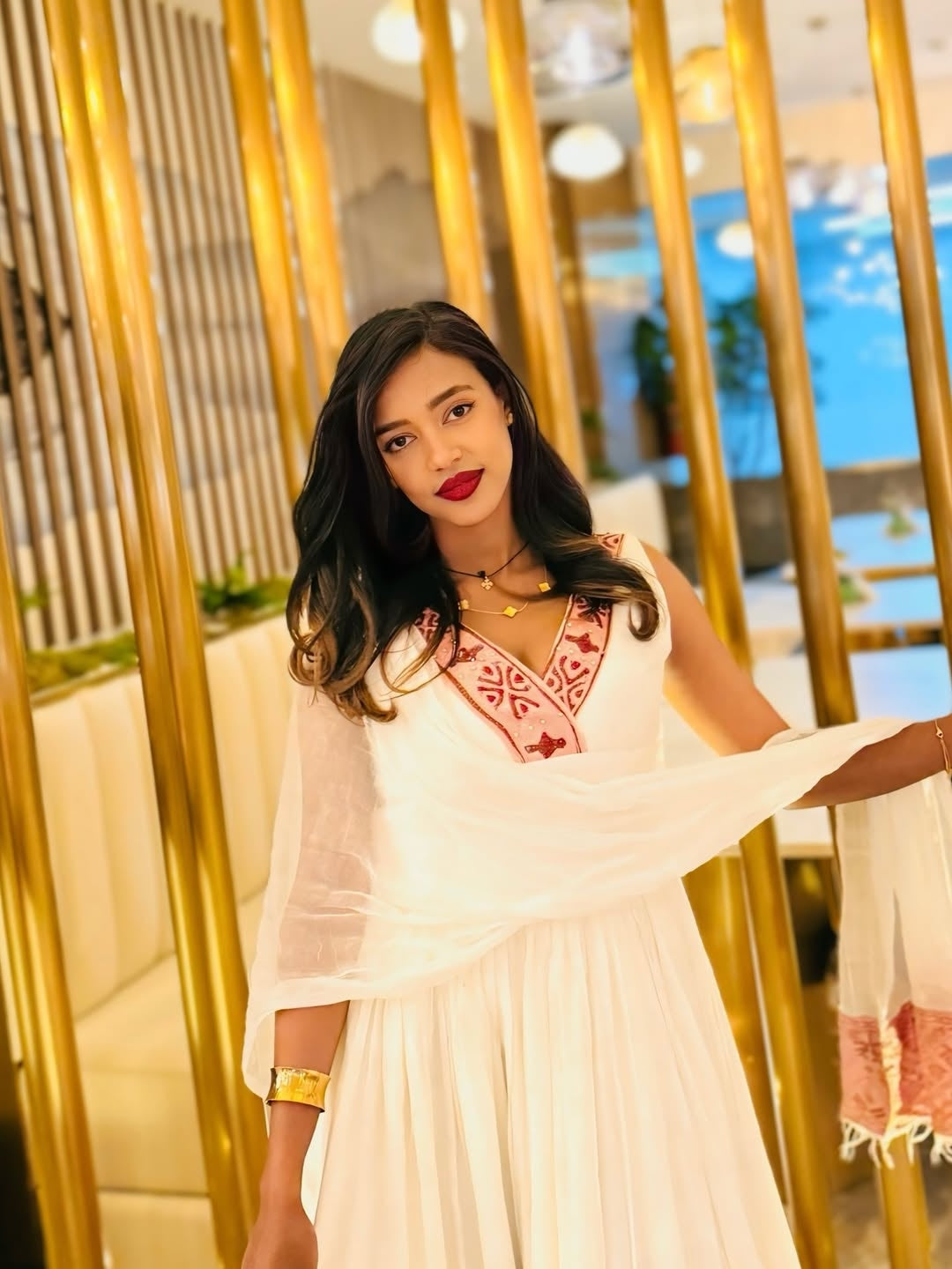 Woman in a white Ethiopian dress with red embroidery standing in a decorated indoor setting.