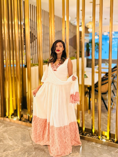 Woman in a white and pink Ethiopian dress standing in a modern indoor setting with gold accents.