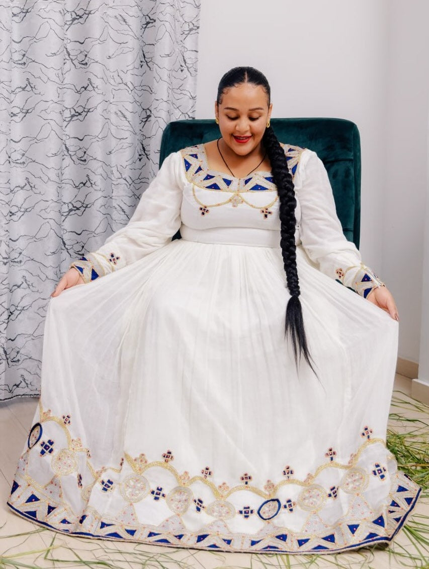 Woman in a white Ethiopian cultural dress with blue and gold embroidery sitting on a chair.