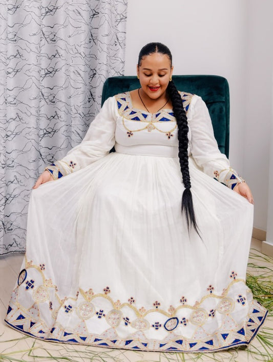 Woman in a white Ethiopian cultural dress with blue and gold embroidery sitting on a chair.
