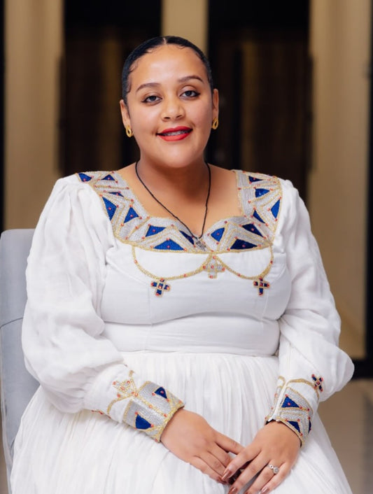 Woman wearing a white Ethiopian Cultural   dress with blue and gold embroidery sitting on a couch.