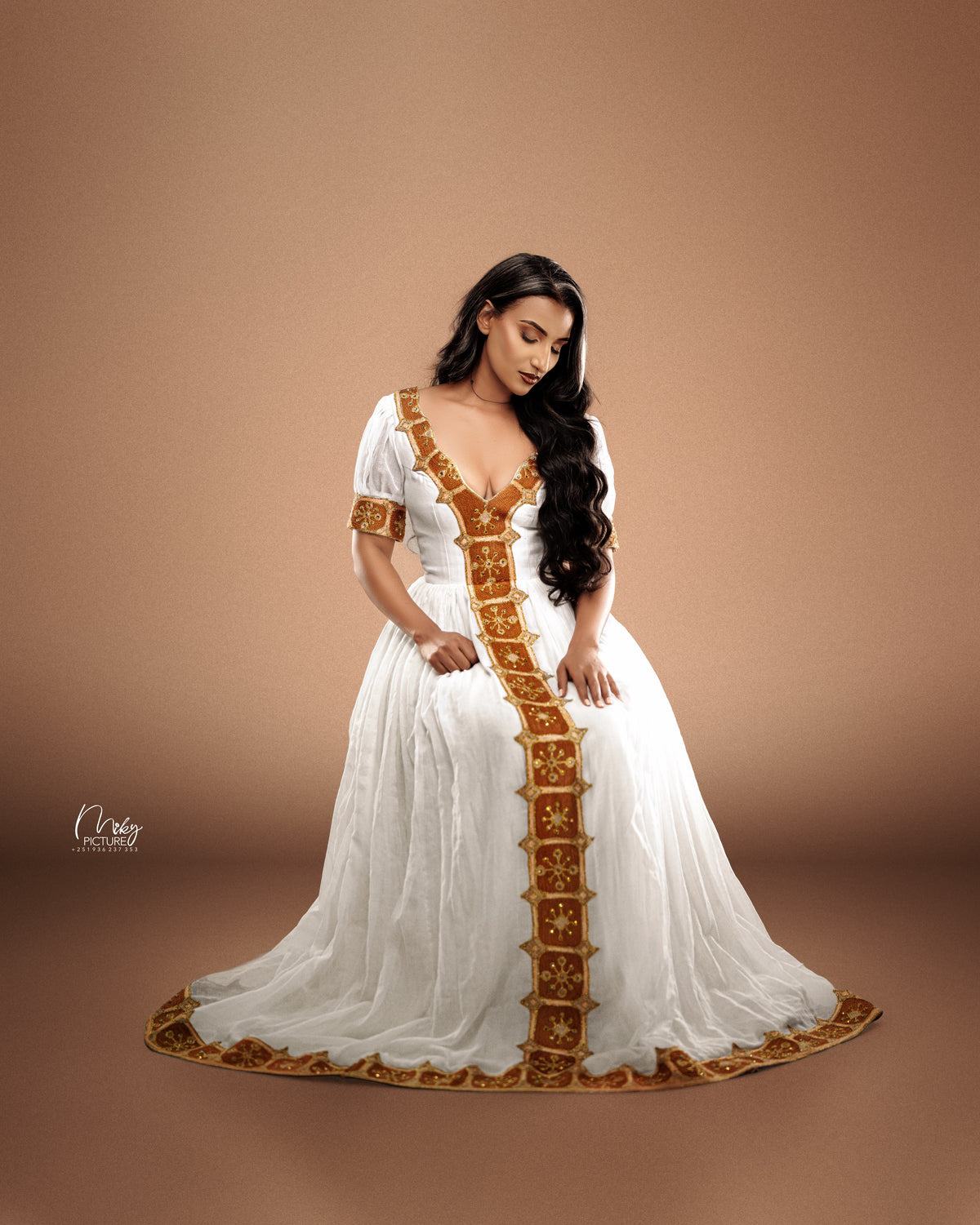 A woman wearing a flowing white traditional Ethiopian national dresses with ornate gold embroidery, posed against a solid warm brown studio background.