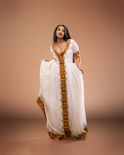 A woman modeling a white Ethiopian national dresses with gold embroidered borders and a deep V-neckline, set against a solid warm brown studio backdrop.
